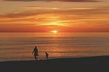 Just above the horizon a blazing orange sky hovers above a beach, where a man and a boy walk, the boy lagging or following as he would.