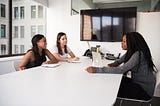 Three women engaged in discussion sitting across a table from each other.