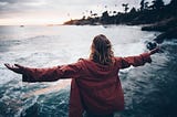 White person with medium-length brown hair in a red hooded jacket, arms outstretched in openness as they look out into the cove of an ocean side.