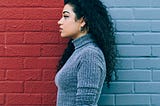 A woman with curly dark hair stands in profile against a wall split into red and blue sections, wearing a grey long-sleeve top and black pants.