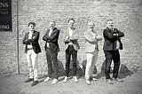 Black and white photo of five men in sport jackets with their arms crossed.