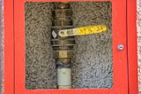 Photo of metallic gas pipe with a valve, in a red security box, the box center has a glass and therefore is transparent, so the valve is visible,