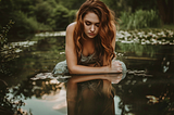 Photo of a woman with long hair, looking at her own reflection at a pond. AI image created by Henrique Centieiro and Bee Lee on MidJourney.