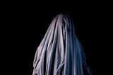 The top half of a figure, probably a woman’s, with its back to the camera, shrouded by a veil or cloth, against a black background