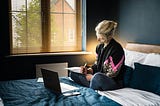 A woman sitting on her bed journaling, with a laptop in front of her in a dark blue bedroom.