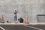 Woman in dress and straw hat with back to camera stares at tall concrete wall. Orange traffic cones and fencing nearby.