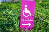 A pink sign on some grass, with a wheelchair symbol, the words “step free route” and an arrow to the right.