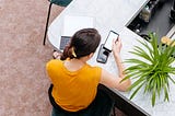 An overhead shot of a woman wearing a yellow shirt, sitting with various gadgets, and using her smartphone.