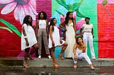 6 Black women dressed in varying shades of white posed against a brick wall painted in colorful flowers
