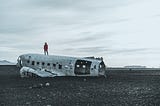 A figure stands on the broken husk of a plane in the middle of a gravel field.