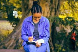 a balck woman dressed in bright blue sits by a tree, writing in her hournal.