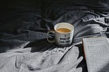 photo of a coffee mug that reads “I woke up like this #tired.” The mug is sitting on gray sheets beside an open book.