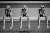 three skeletons on shelf doing See No Evil, Hear No Evil, Speak No Evil