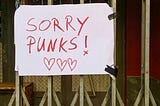 Handwritten poster taped to metal bars of a gate. Poster reads, “Sorry Punks” with three lovehearts drawn underneath.