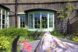 A view across a small overgrown garden to a bow window and a grey pebbledash wall with plants growing up it. In the foreground the end of a garden sofa with a pair of feet in stripy red socks (my own) and a black tail (my cat’s)