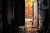 A chair sits in the open door at the end of a dark enclosed alley that promises light outside.