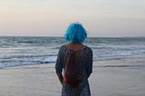 A woman with blue hair staring out into the ocean admiring its calm scenic beauty.