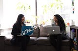 Two women of color smile at each other with open laptops.