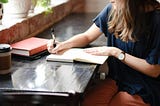Woman sitting at a wooden desk and writing in a notebook, only she’s millennial-pink levels of cool and polished.
