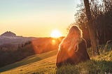 color photograph of someone with long blonde hair sitting on a grassy hill watching the sunset