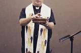 The author wearing Christian clerical attire (white collar, black shirt, and white stole), holding a tray of communion bread