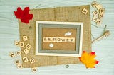 the word empower on tiles in a frame. Laying on a piece of burlap surrounded by extra tiles, and leaves.