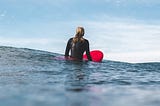 A female surfer clad in a wetsuit, sitting in the ocean on her red surfboard, waiting for waves