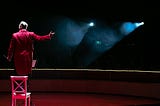 A ringmaster in a circus standing on a chair introducing the next event.