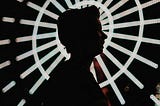 Person silhouetted against the lit spokes of a ferris wheel, as if the light is emanating from them