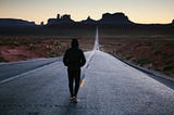A hooded figure walks along the yellow, dashed line in the center of a road going up into the mountains on the horizon.