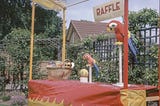 A stall with a sign saying ‘raffle’ on it. The stall is covered in red and yellow material and is situated in a garden or other green space. There is a basket with what looks like small prizes in sitting on the stall as well as two brightly coloured statues of parrots on perches and a yellow ball.