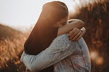 Two young people embrace in a field of grass beneath the glow of late afternoon sun.