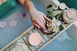 Uma cena serena de um spa mostra uma mão descansando em um suporte de madeira sobre uma banheira. No suporte, encontram-se elementos típicos de uma experiência de spa, como um pote de sais de banho cor-de-rosa, uma vela aromática suave, rodelas de pepino fresco, e flores brancas delicadas. A água azulada da banheira está adornada com pétalas de flores, sugerindo um ambiente relaxante e luxuoso, ideal para um momento de autocuidado profundo.