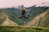 Person with long hair in athletic wear photographed jumping up in a celebratory fashion on top of a mountain.