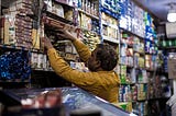 A kirana store vendor pulling out the requested item from his shelf filled with different household items