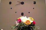 A unique wall clock. In front of it, is a table and chairs with a bouquet of flowers.