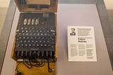 Museum display shows Enigma coding machine, black with keys like a typewriter, on left and info card on right.