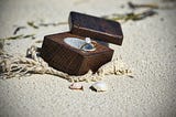 A little wooden box resting on a sandy beach with a beautiful diamond ring inside.