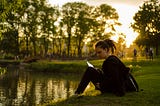 Girl sitting on a grass patch with a book by a body of water