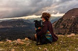 A woman hugging her dog and watching a storm roll in while her dog checks for activity behind them.