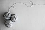 Three balls of light grey yarn with a singular thread looping at the top on a grey background