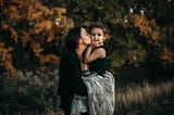 Portrait of mother kissing her young child in front of autumn trees