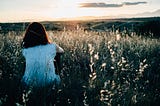 Female with short dark brown hair sitting in field in hilled area with sunset in the background.