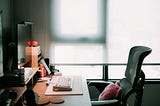 A softly lit home office setup with a modern ergonomic chair, wooden desk, computer keyboard, and monitor. Warm light filters through a frosted window in the background, creating a calm, focused atmosphere. A pink cushion rests on the chair, adding a touch of comfort and personality to the workspace.