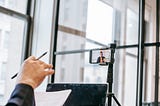 Fotografia de uma pessoa em um ambiente de escritório, participando de uma videoconferência ou gravação usando um smartphone montado em um tripé. A câmera do smartphone está voltada para a pessoa, que aparece na tela do dispositivo enquanto fala ou apresenta algo. A mão de outra pessoa, segurando uma caneta e um pedaço de papel, também aparece em primeiro plano, sugerindo que a pessoa está envolvida na conferência, talvez como espectadora ou participante