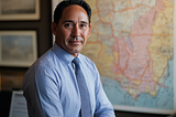 A man in a blue shirt and tie stands in front of a map with areas highlighted in yellow and red.