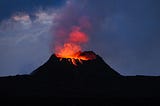 A volcano erupts at twilight, lava is dripping down the sillouetted volcano, and red smoke rises from the cone.