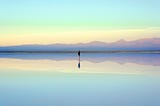 Single figure standing on water which is reflecting a clear sky