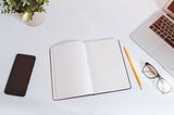 A phone, notebook, pencil, glasses, plant, and laptop on a table.