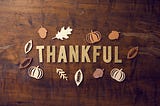 The word “Thankful” spelled out on a wooden surface, surrounded by paper cutouts of leaves, acorns, and pumpkins